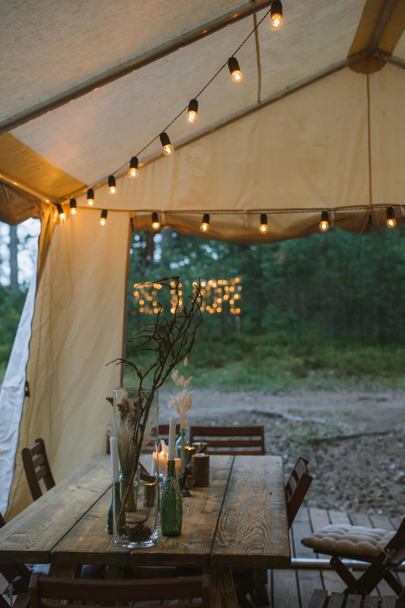 An open tent with string lights over a rustic table set for a gathering in the woods